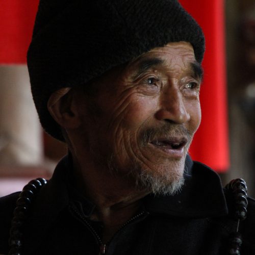 A smiling older Tibetan man wearing black prayer beads.