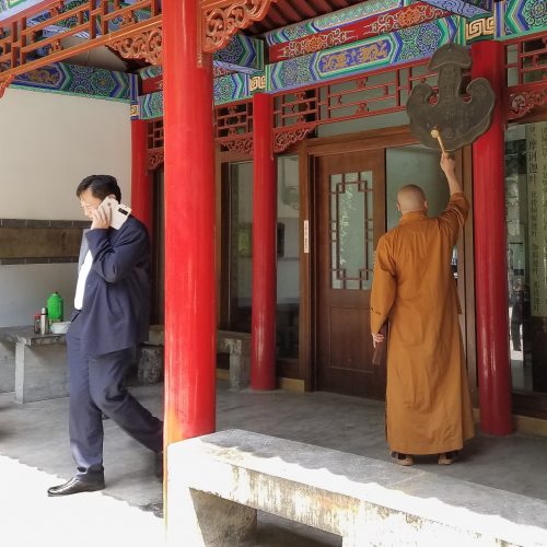A man in a suit talking on a cell phone while behind him a man wearing monk's attire rings a gong.