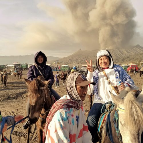 Two women on horseback smiling, one making a peace sign, with an active smoking volcano in the background.