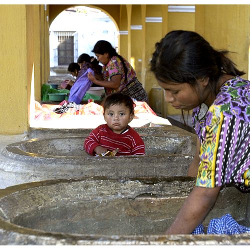 Women working on washing clothes while a young boy stares curiously at the camera.