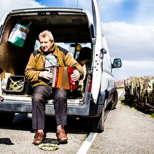 A man sitting in the back of his van with an accordion while his dog stands next to him.