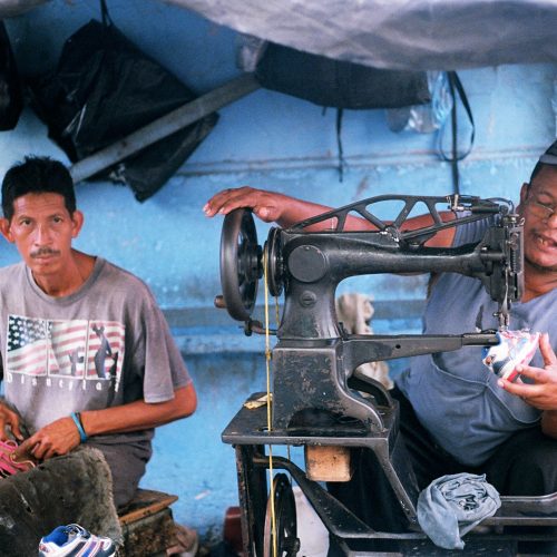 Two men working on making shoes, one using a sewing machine.