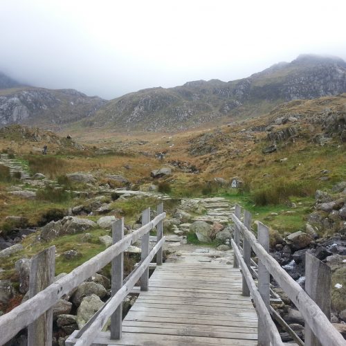 A wooden bridge leading to grassy hills and mountains.