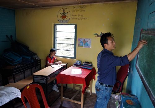 A young Indian boy watches a teacher write on a chalkboard.